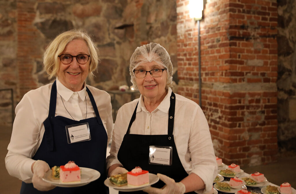 Kaksi iloista naista tummansinissä essuissa. He pitävät käsissään lautasta, jossa leivos ja piirakkaa. 