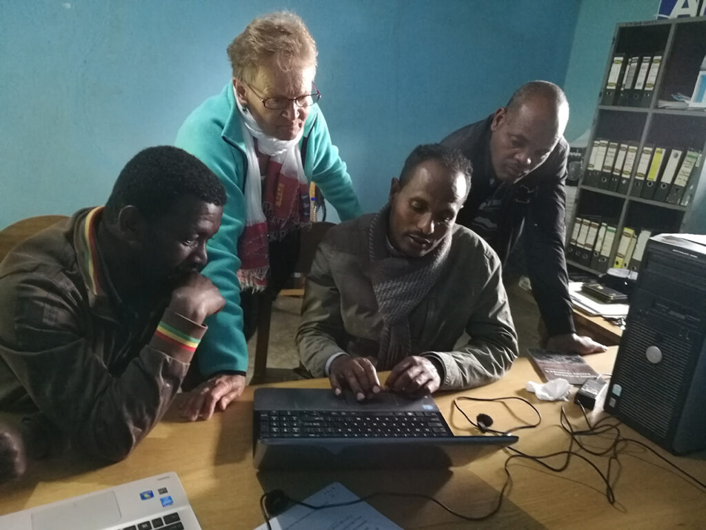 Neljä etiopialaista miestä ja yksi vaalea suomalaisnainen kurottava kohti tietokonetta huoneessa.