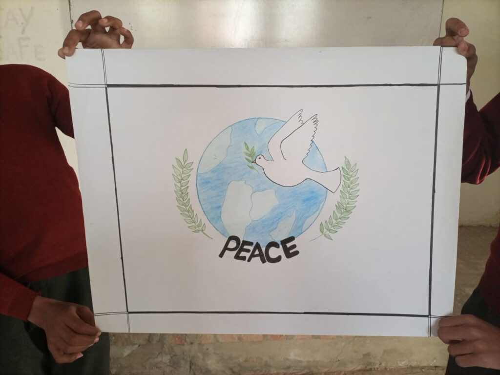 Kuvassa näkyy kahden henkilön välissään pitämä piirustus. Piirustuksessa on valkoisella taustalla maapallo ja rauhankyyhkynen. Kuvan alhaalla lukee peace eli suomeksi rauha.