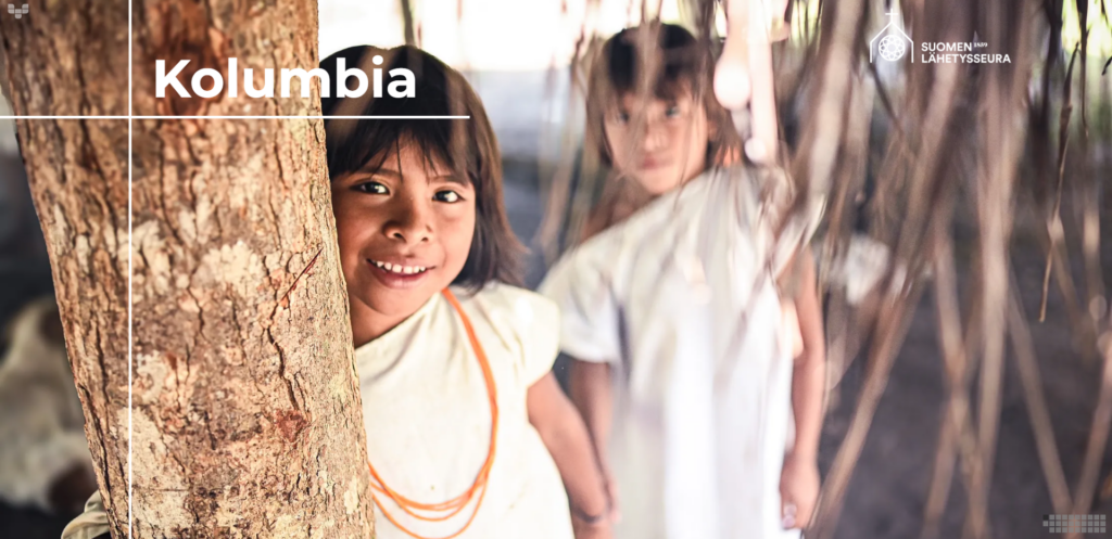 Kaksi kogi-alkuperäiskansaan kuuluvaa lasta puun takana, kuvan päällä lukee Kolumbia