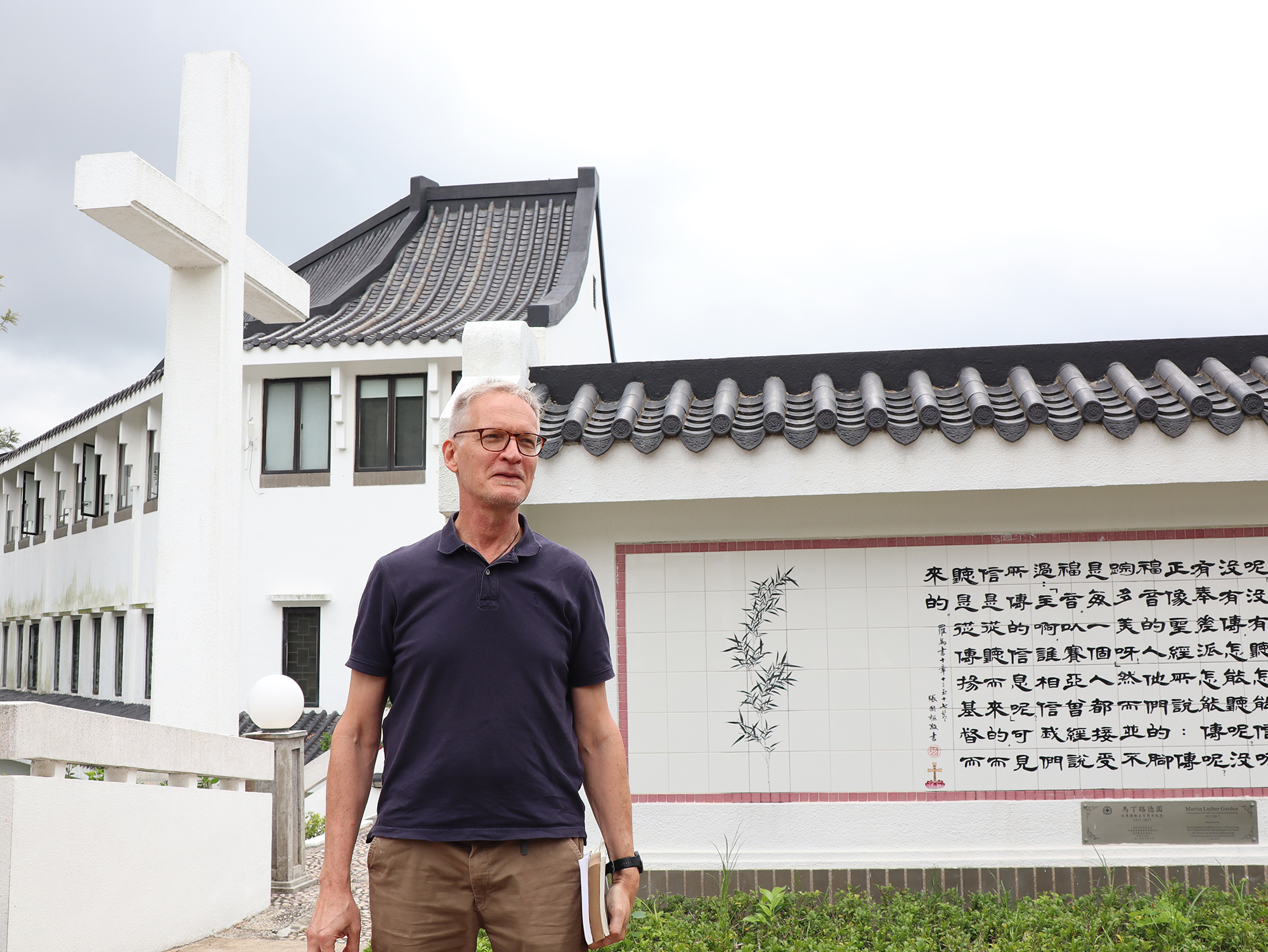 Jukka Helle seisoo seminaarin pihalla kädessään kirjapino. Taustalla näkyy iso risti ja valkoinen kiinalaisin kirjaimin kaiverrettu kivitaulu.